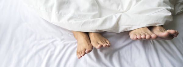 Feet of couple sleeping side by side in comfortable bed. Close up of feet in a bed under white blanket. Bare feet of a man and a woman peeking out from under the cover.Top view with background copy space.
