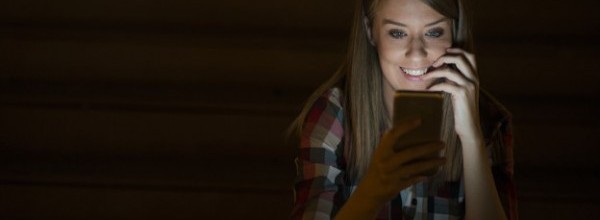 Gorgeous young woman standing with mobile phone at night street outdoors, female caucasian student reading text messages on her cell phone with reflected on her face screen light, blank screen phone